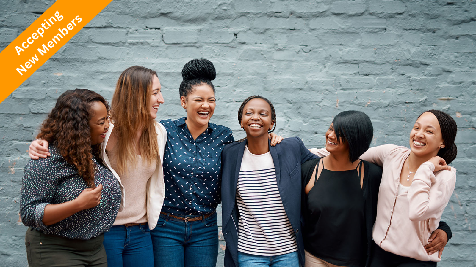 Portrait of a diverse group of young women smiling, laughing, and standing together against a gray wall outside.
